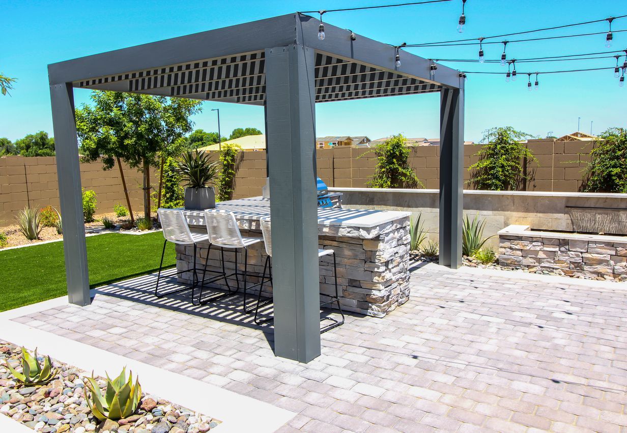 Outdoor bar with stone facade under gray pergola on a paved patio, with a green lawn and plants.