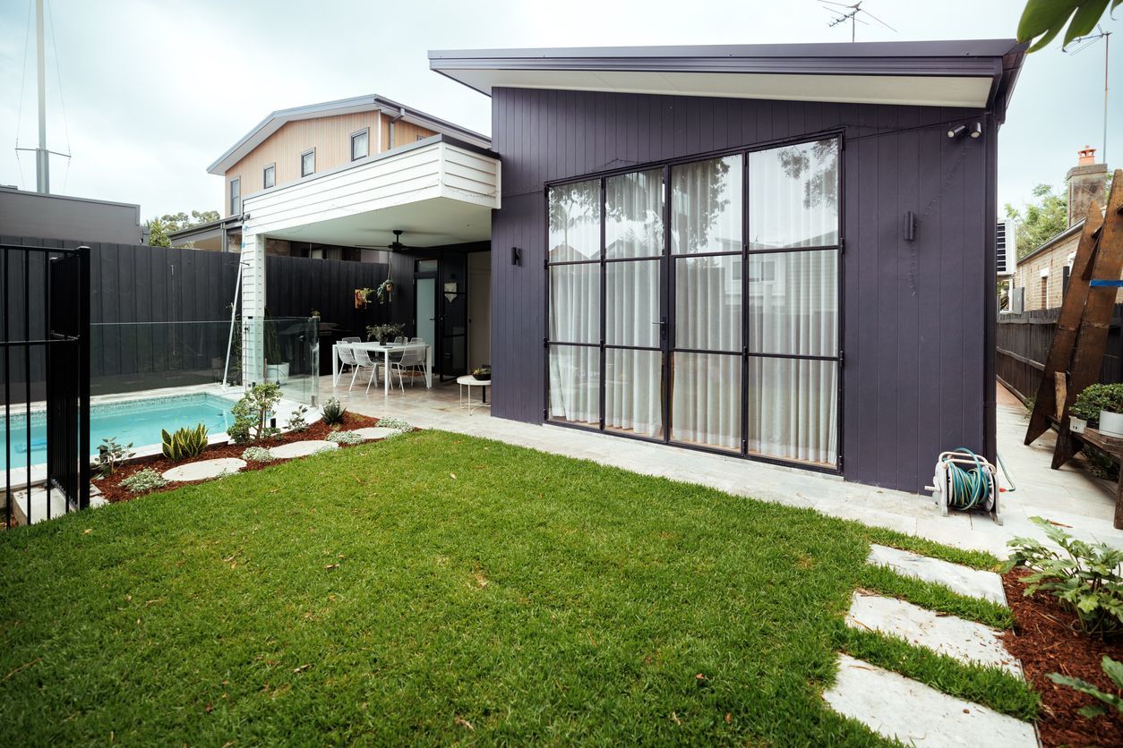 Modern home exterior with dark siding, large windows, patio, pool, and green lawn.