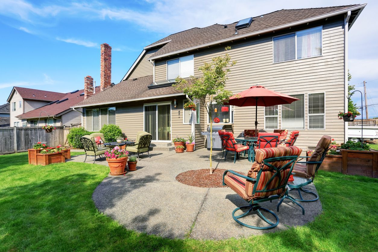 Backyard patio with seating, grill, and umbrella, adjacent to a two-story house with green lawn.