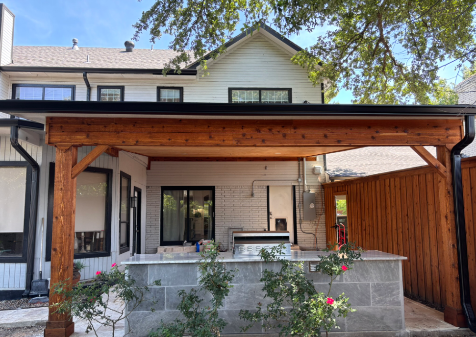 Wooden patio cover attached to a white house with a stone counter and a grill.