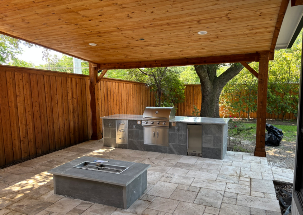 Outdoor kitchen patio with overhead wooden pergola, grill, fire pit, and fence.