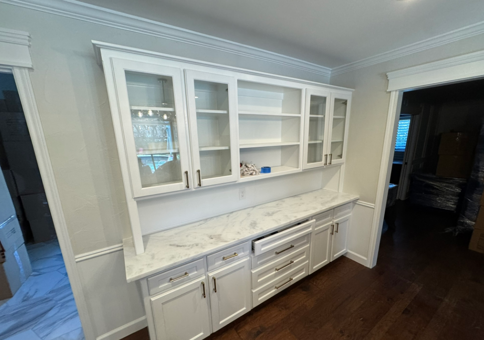 White built-in kitchen cabinet with glass-front uppers and marble countertop. Dark wood floor.