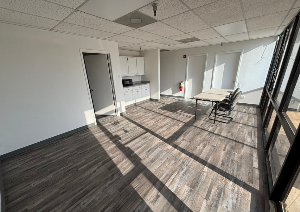 Empty office space with wood-look flooring, kitchenette, meeting table, large windows, and sunlight.