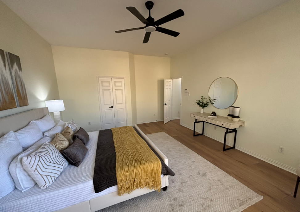 Bedroom with bed, rug, vanity, door, and ceiling fan; beige walls, natural light.