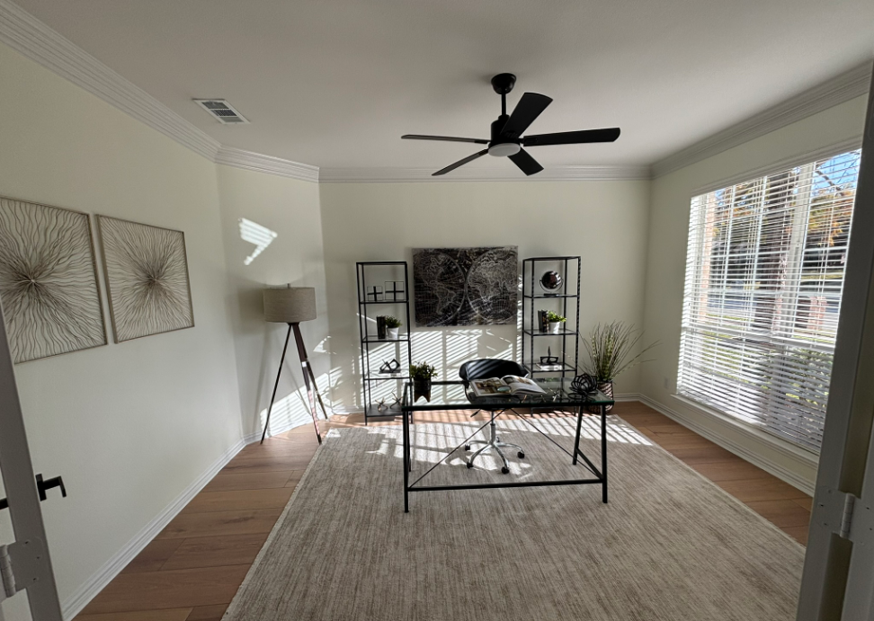 Office with desk, shelves, art, and a large window with blinds. Neutral color scheme and hardwood floors.