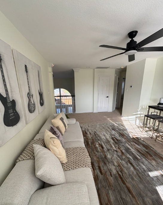 Living room with a large sofa, guitar art, and a wood-look rug. Black ceiling fan and doorway.