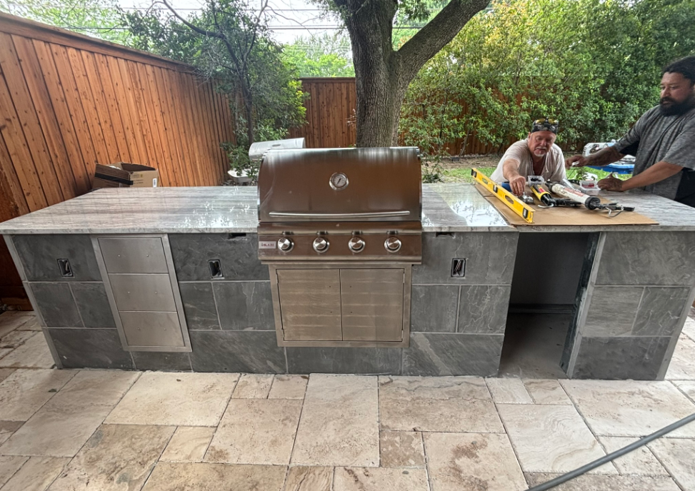 Outdoor kitchen with a built-in grill, two people working, gray stone and countertop.