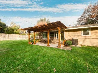 Backyard with green grass, wooden pergola over patio, beige house, and fence.