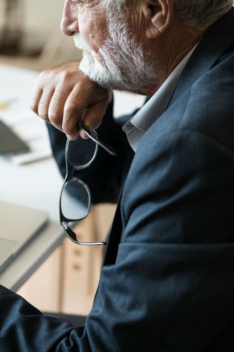 A person in a dark suit rests their chin on their hand while holding a pair of glasses at a desk.