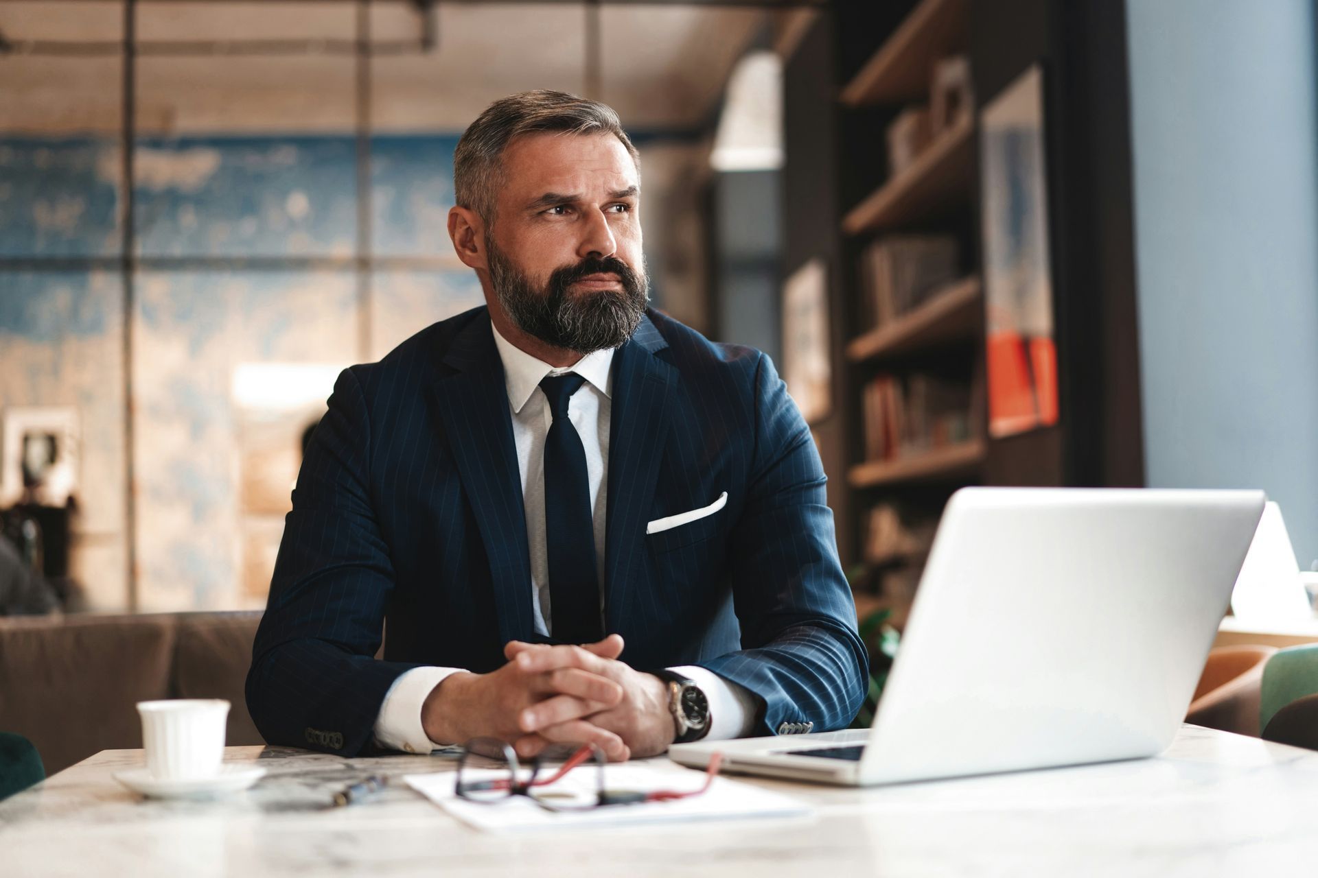 A professional in a suit and tie sits at a table with a laptop, coffee, and glasses, looking away thoughtfully.