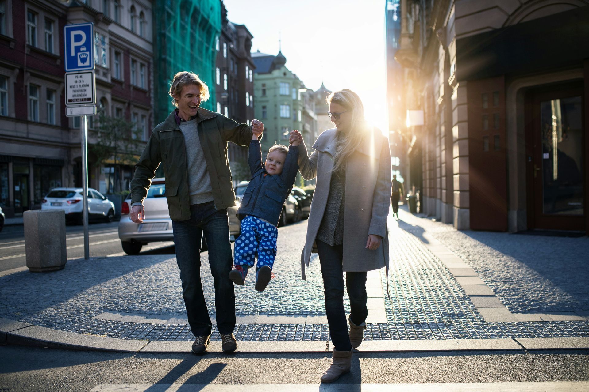 A family walking on a sunny city street, swinging their child between them, backlit by the sun.