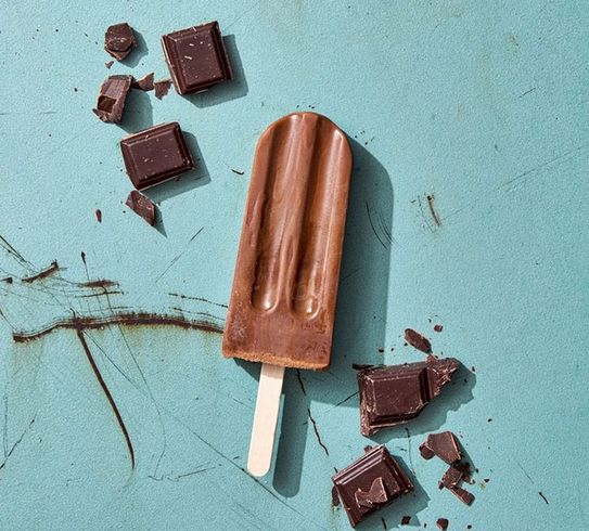 A chocolate popsicle is sitting on a blue surface next to pieces of chocolate.