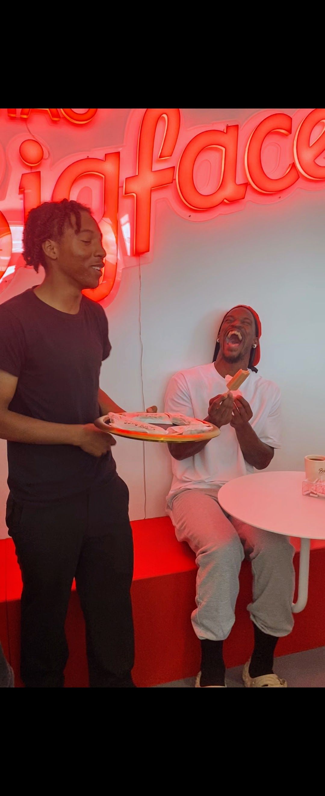 Two people in a cafe. One holds a tray, the other sits at a pink table. Red and pink decor.