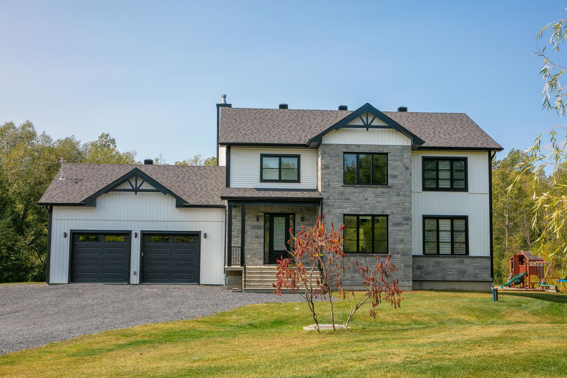 Maison à deux étages avec façade en pierre grise, revêtement blanc, garnitures noires et toit gris, située sur une pelouse verte.