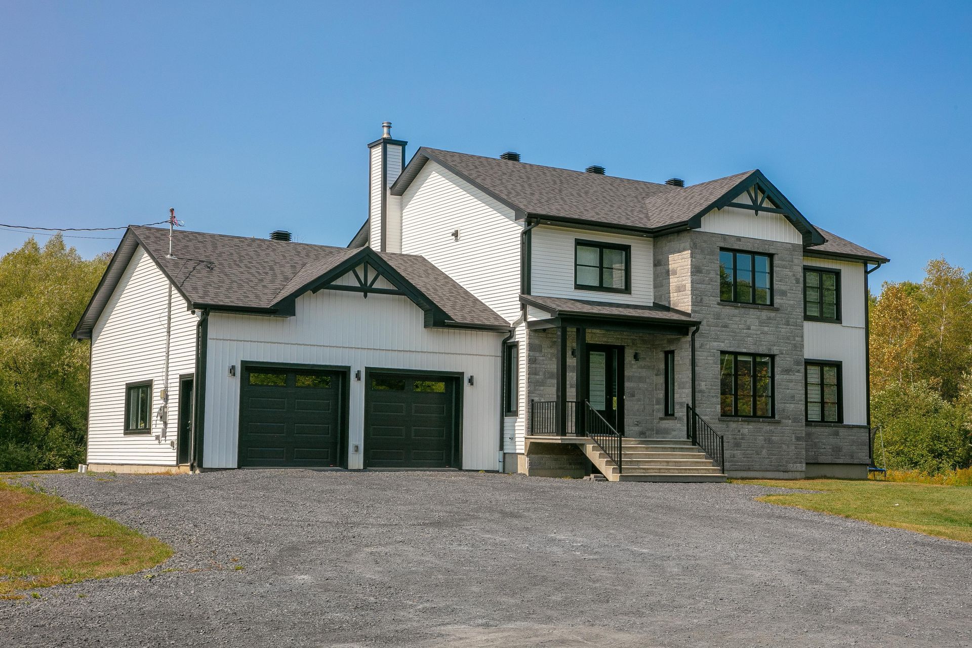 Maison à deux étages avec revêtement blanc, façade en pierre et portes de garage noires sur une allée de gravier sous un ciel bleu.