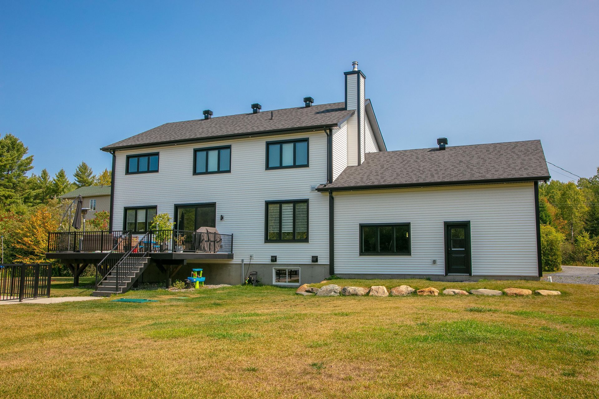 Maison blanche à deux étages avec des garnitures noires, une terrasse et une cour sous un ciel bleu.