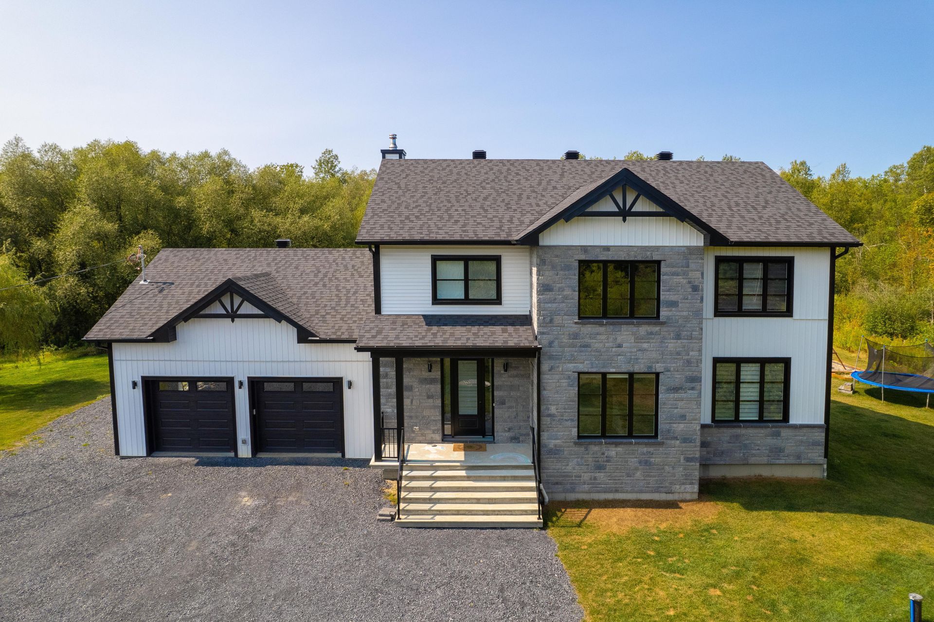 Maison à deux étages avec revêtement blanc, façade en pierre grise, toit noir et garage pour deux voitures; allée sur un terrain gazonné.