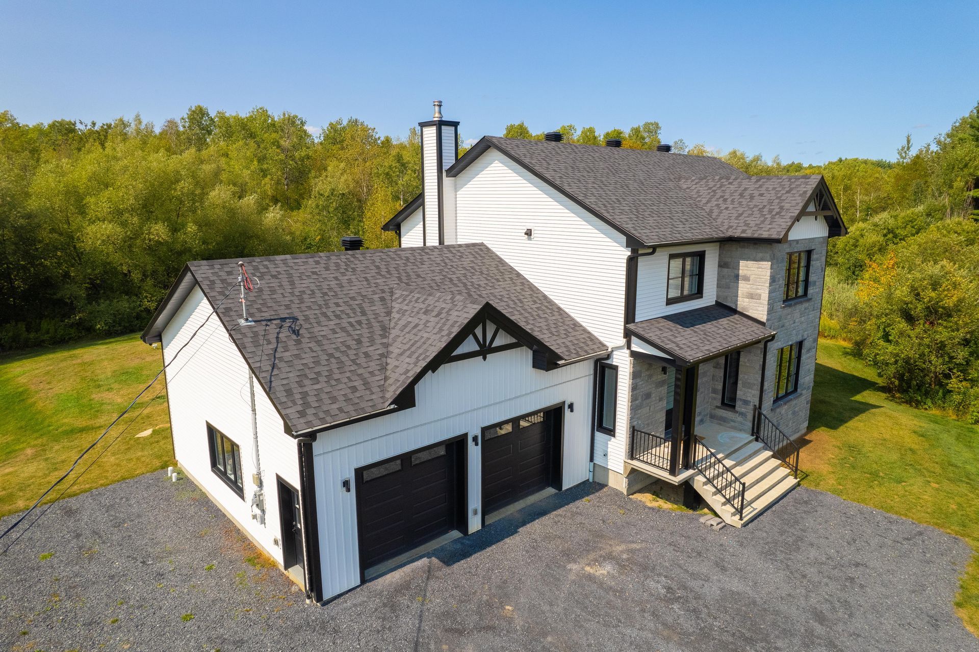 Maison blanche à deux étages avec garniture noire et garage, façade en pierre grise, allée et arbres environnants.