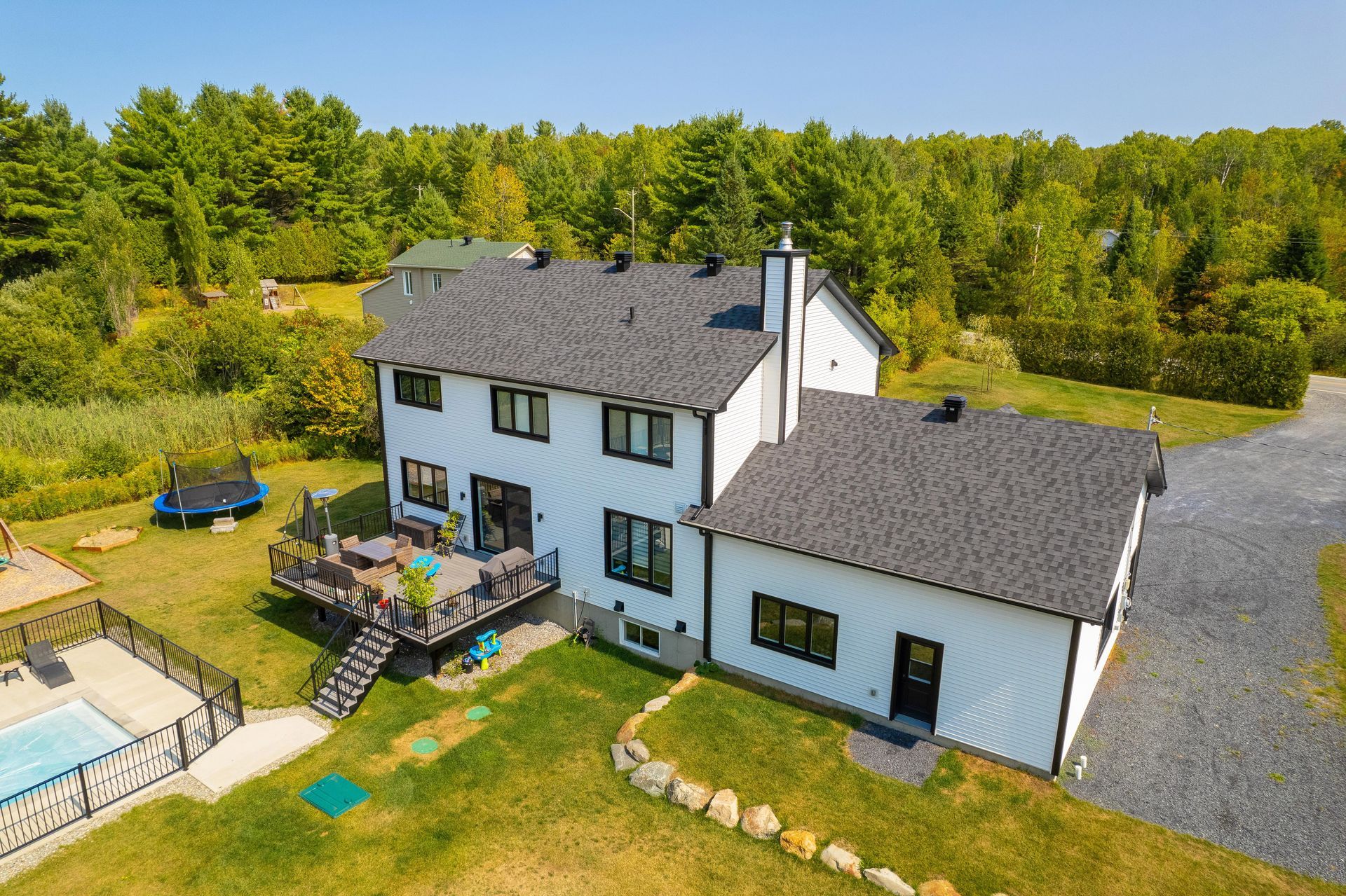 Maison blanche avec garniture noire, cour arrière avec piscine, trampoline et terrasse, entourée d'arbres.