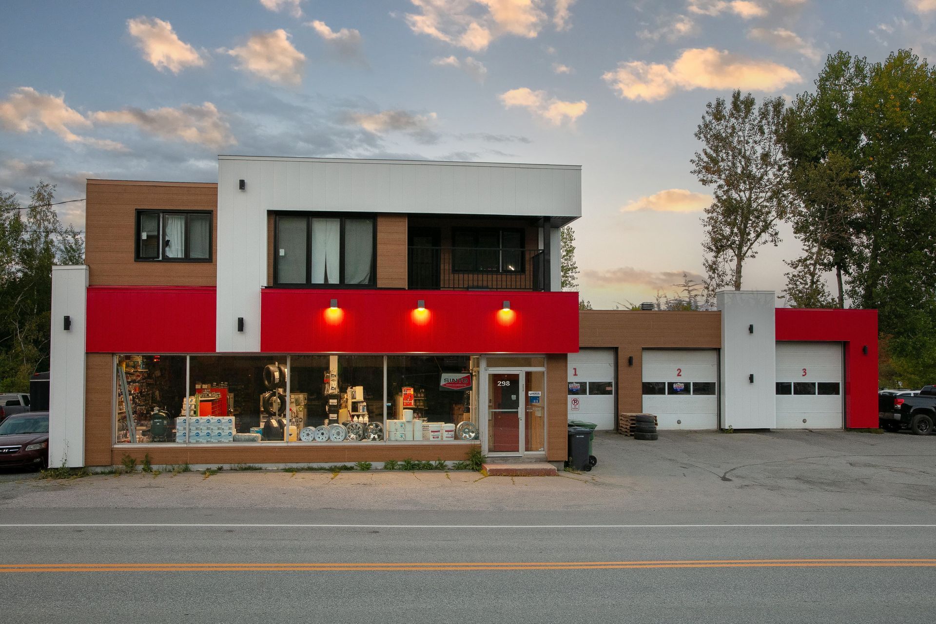 Bâtiment moderne de deux étages aux accents rouges et blancs. Vitrines, portes de garage et rue en façade.