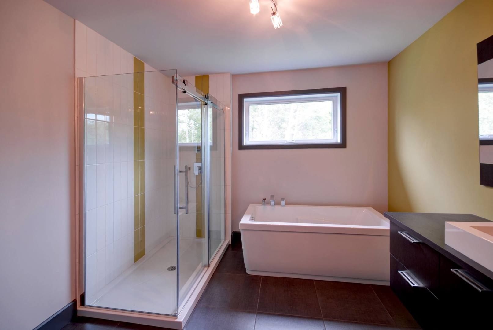 Salle de bain moderne : douche en verre, baignoire blanche, vanité foncée, sol en carrelage noir, fenêtre, murs jaune clair et blancs.