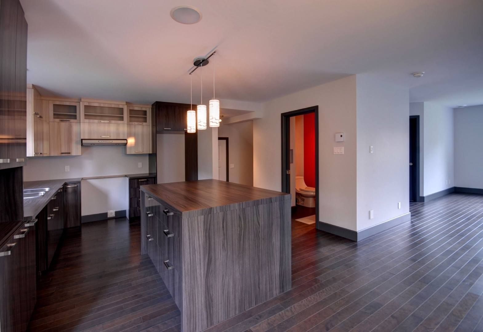 Cuisine moderne avec îlot, armoires et planchers en bois foncé, contrastant avec des murs et des comptoirs clairs.