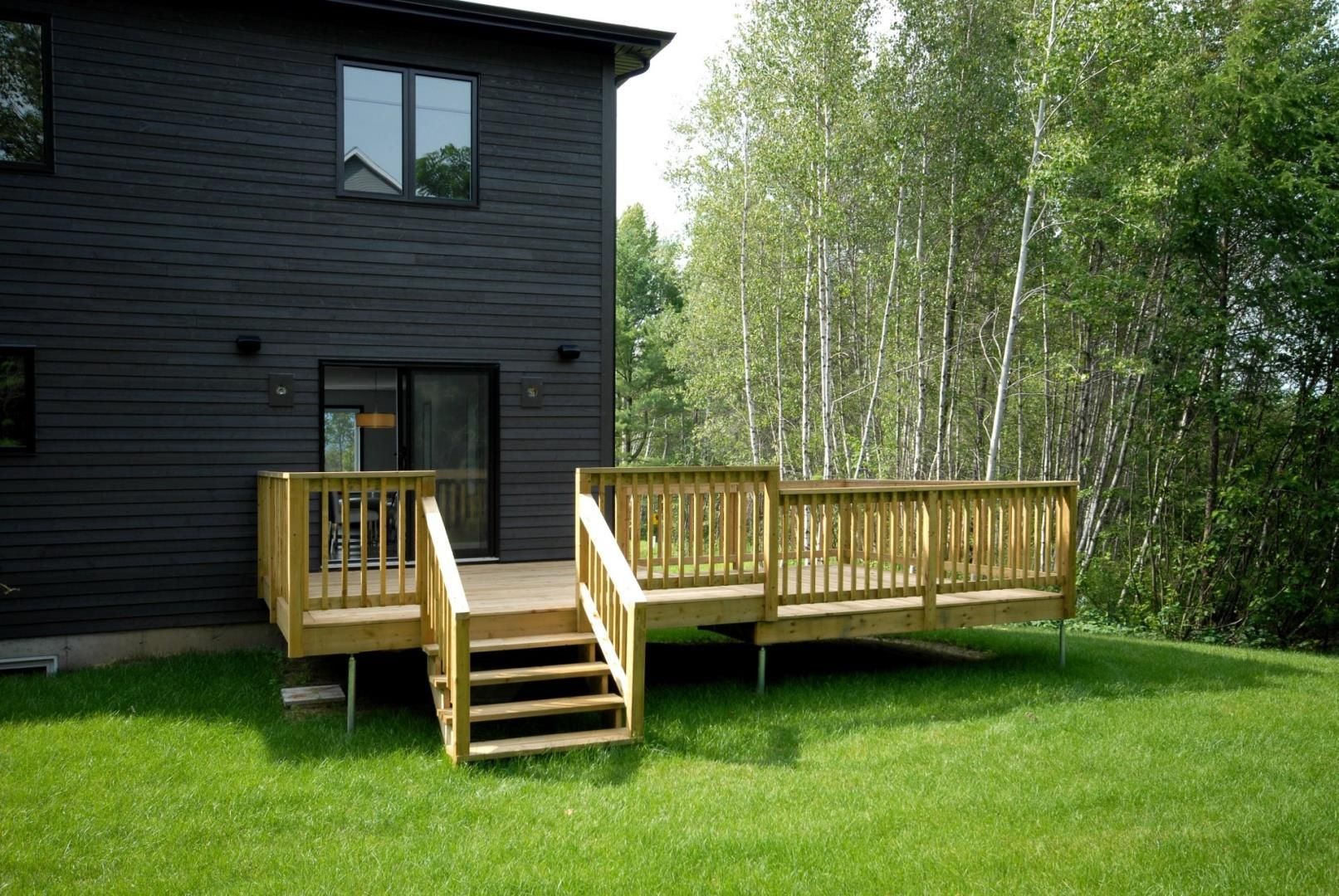 Terrasse en bois attachée à une maison noire, avec des escaliers menant à une cour arrière herbeuse avec des arbres.