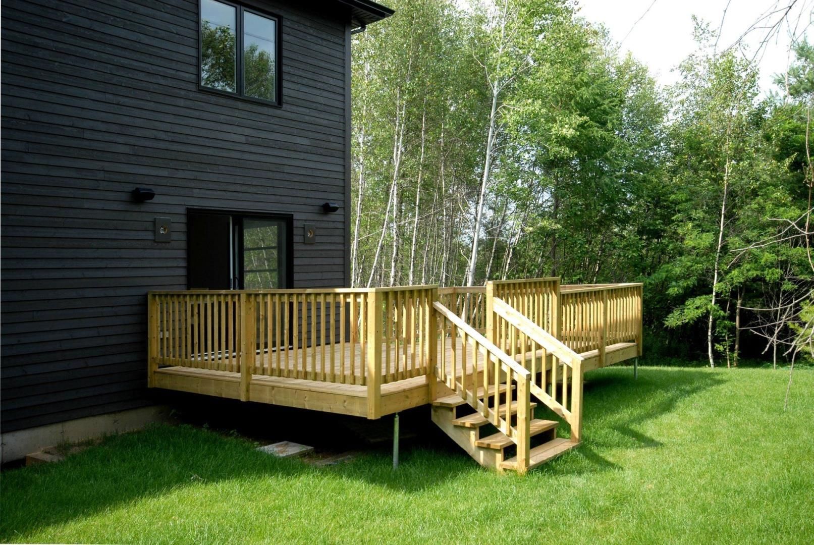 Terrasse en bois attachée à une maison sombre, avec des marches menant à une cour herbeuse et à des arbres.