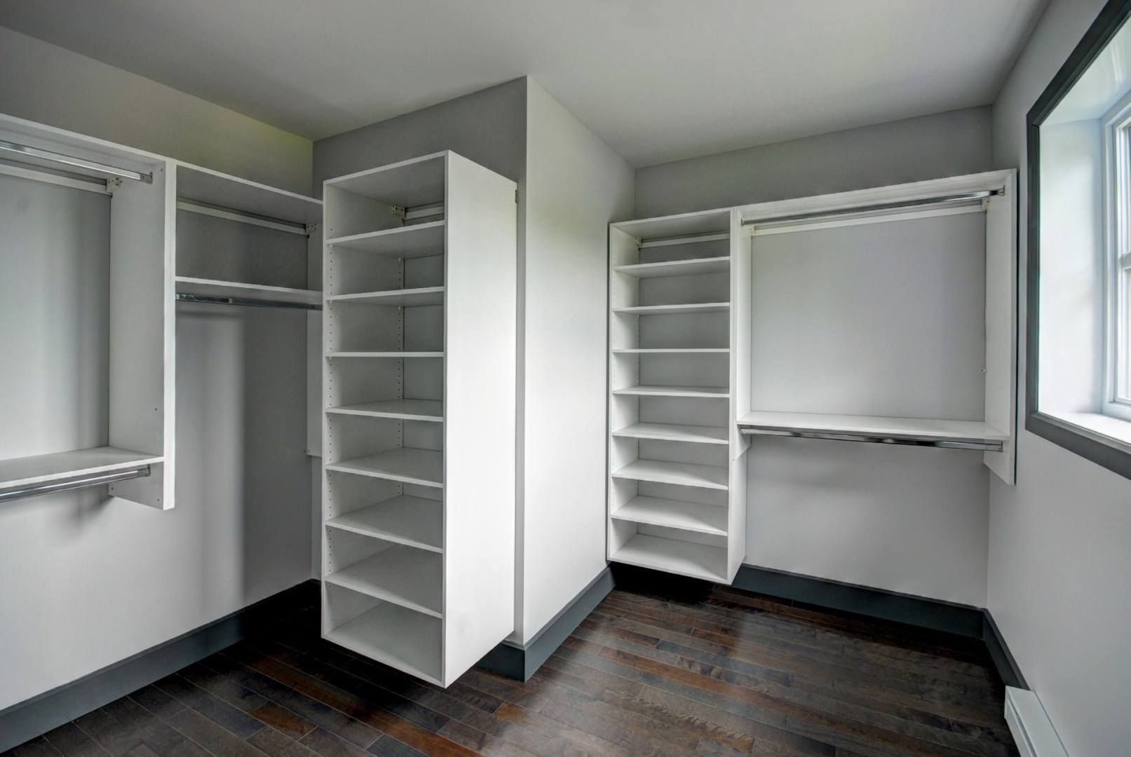 Dressing vide avec étagères blanches et tringles de suspension contre des murs gris, un parquet en bois foncé et une petite fenêtre.