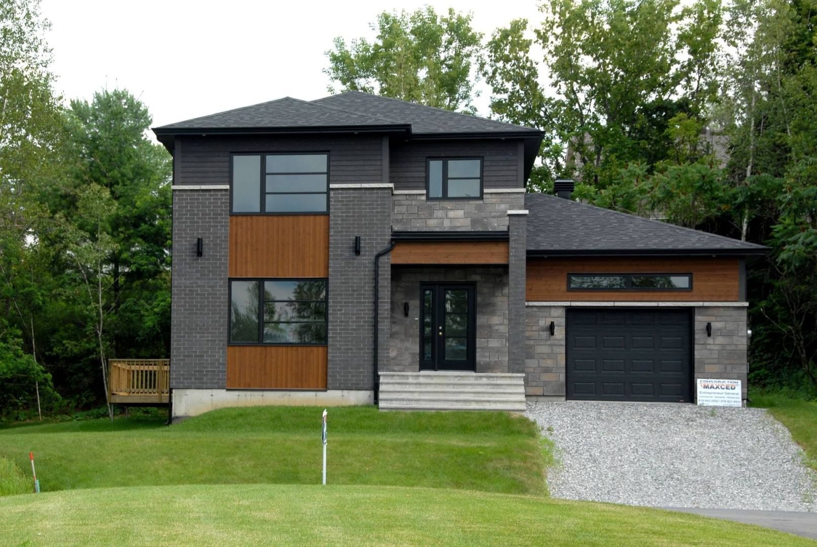 Maison moderne à deux étages avec façade grise et marron, boiseries noires, garage et allée en gravier. Pelouse verte et arbres en arrière-plan.