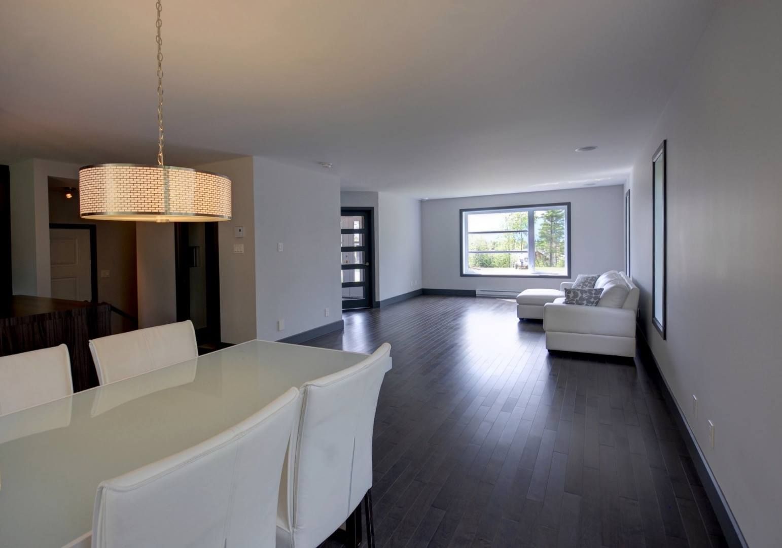 Salle à manger et salon combinés, murs blancs, planchers de bois franc foncé, meubles blancs.