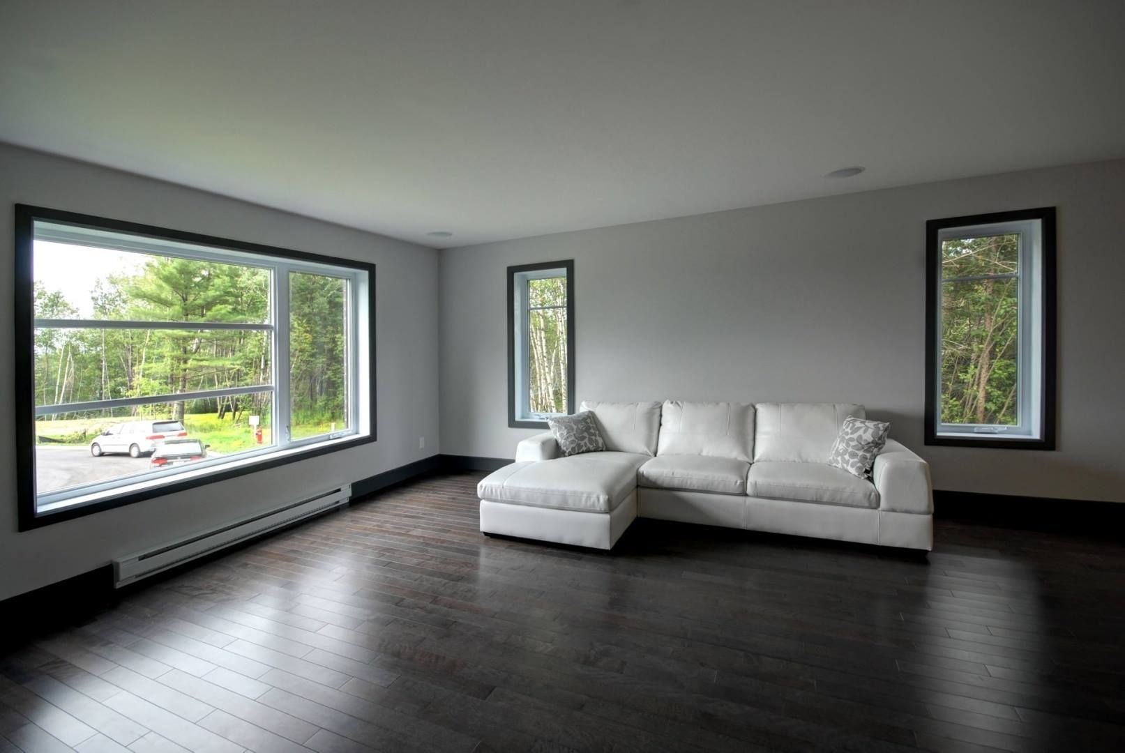 Salon avec canapé blanc, parquet en bois foncé et grandes fenêtres avec vue sur les arbres.