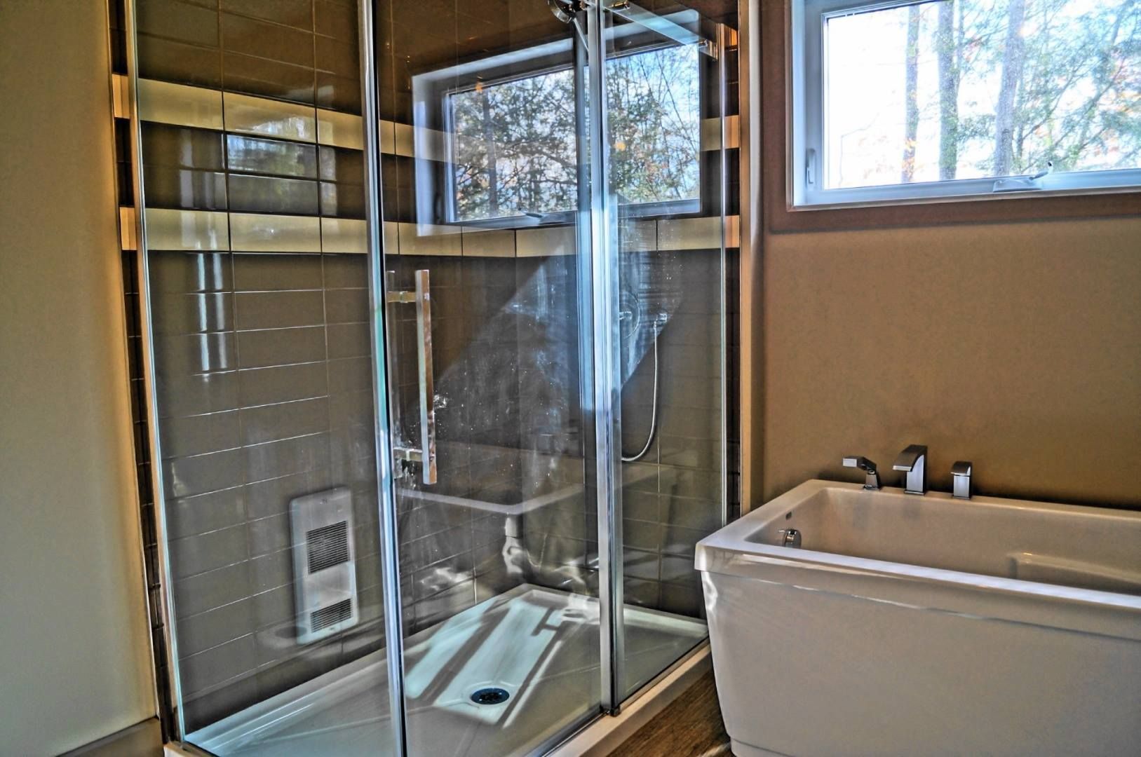 Cabine de douche avec portes vitrées, adjacente à une baignoire blanche, dans une salle de bain avec fenêtre.