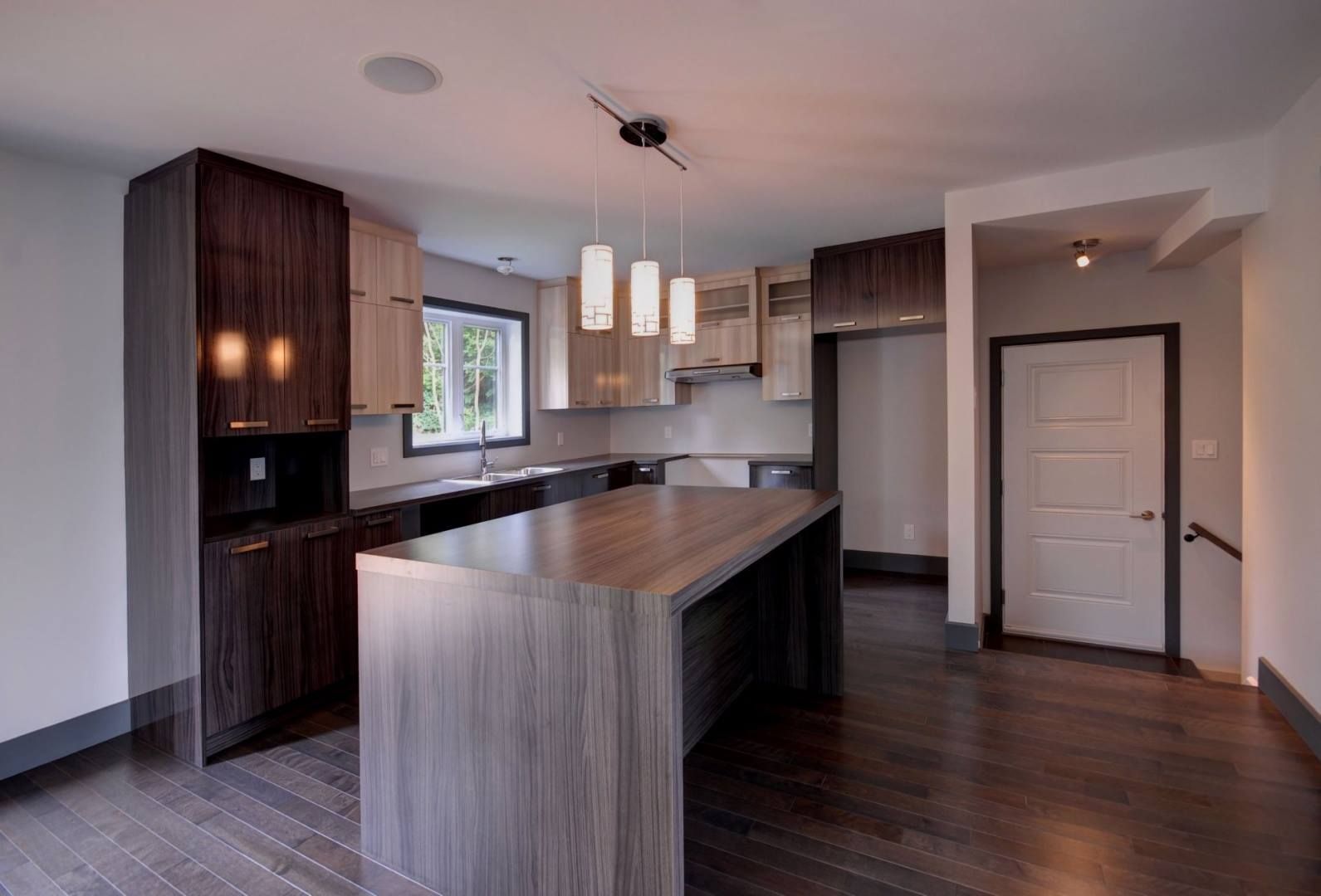 Cuisine moderne avec armoires en bois, îlot central et luminaires suspendus. Parquet en bois foncé.