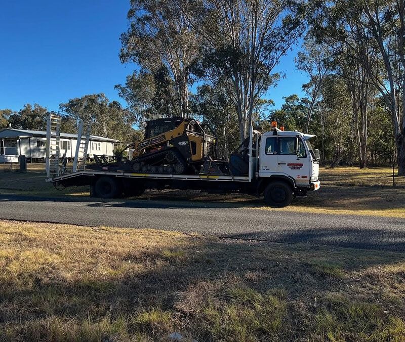 White Flatbed Truck Carrying a Small Bulldozer on a Paved Road — Dudgeon’s Earthworks in Murgon, QLD