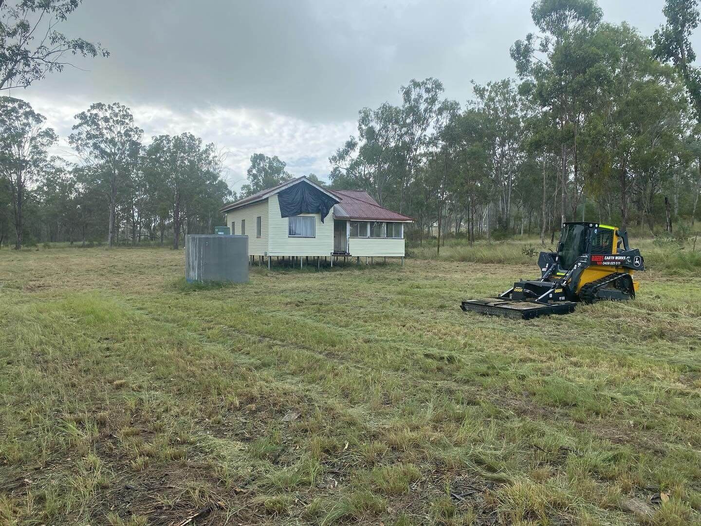 A Skid Steer Mower Cuts Grass Near a Small House and Water Tank, in a Field With Trees — Dudgeon’s Earthworks in Murgon, QLD