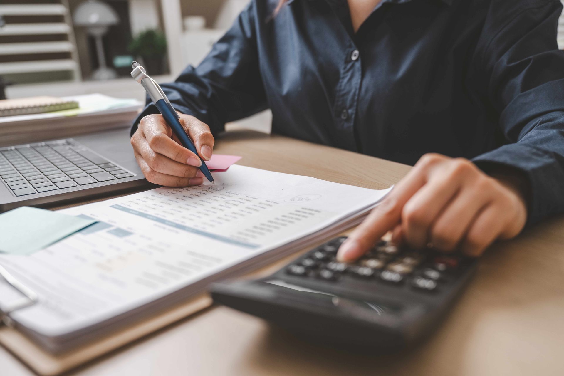 Person using a calculator and pen to review documents at a desk, near a laptop.