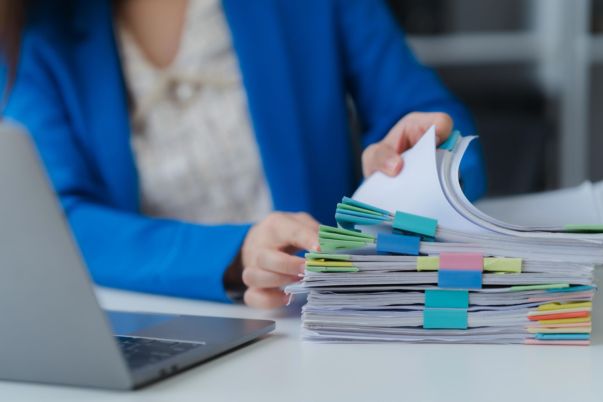 Person in blue jacket sorts papers at a desk with a laptop; documents are organized with colorful tabs.