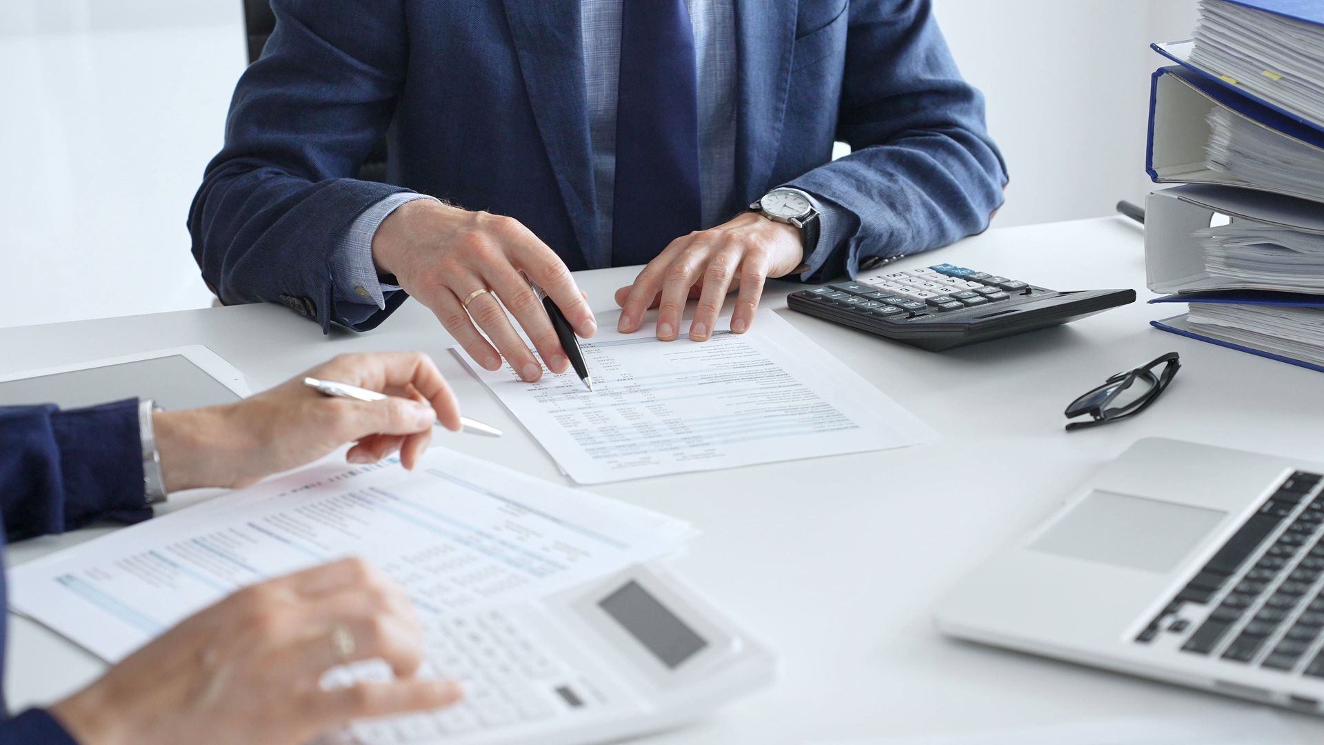 Two people in suits reviewing documents at a desk with calculator and laptop.