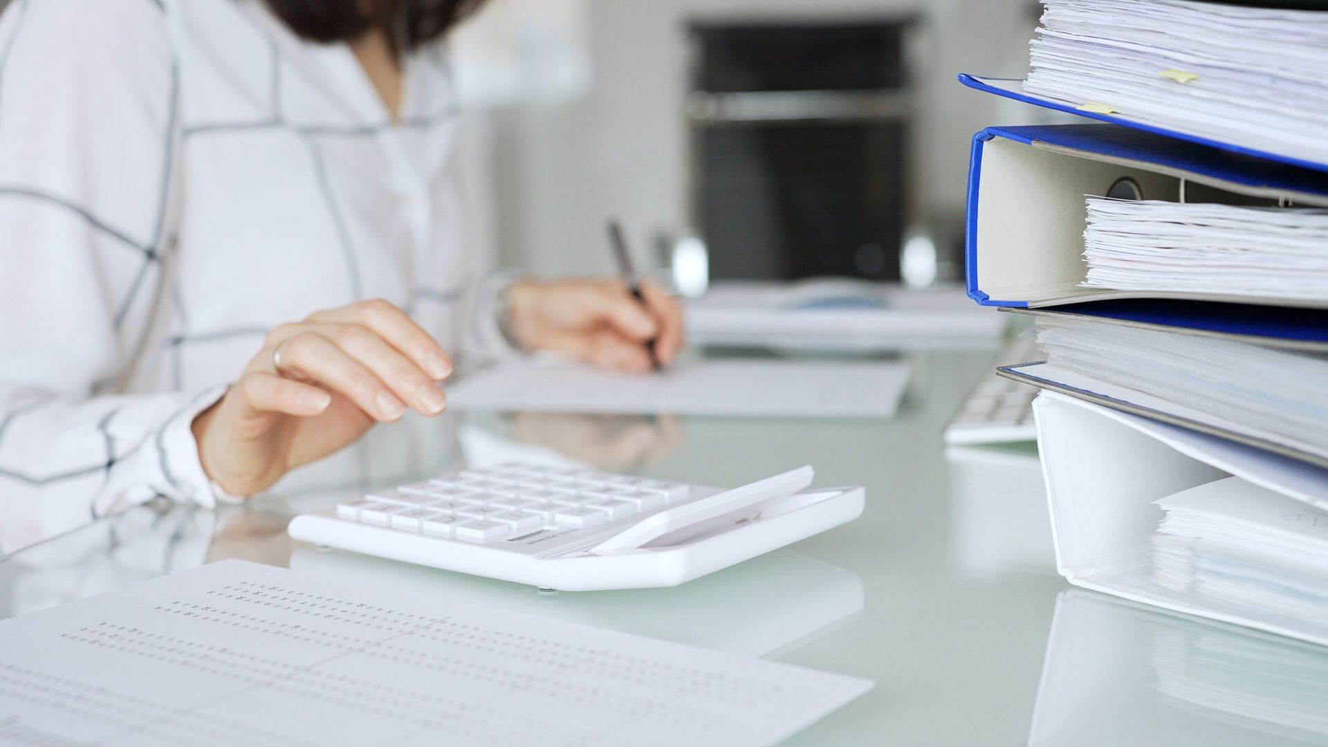 Woman using calculator and writing on paper at a desk, with binders in the background.