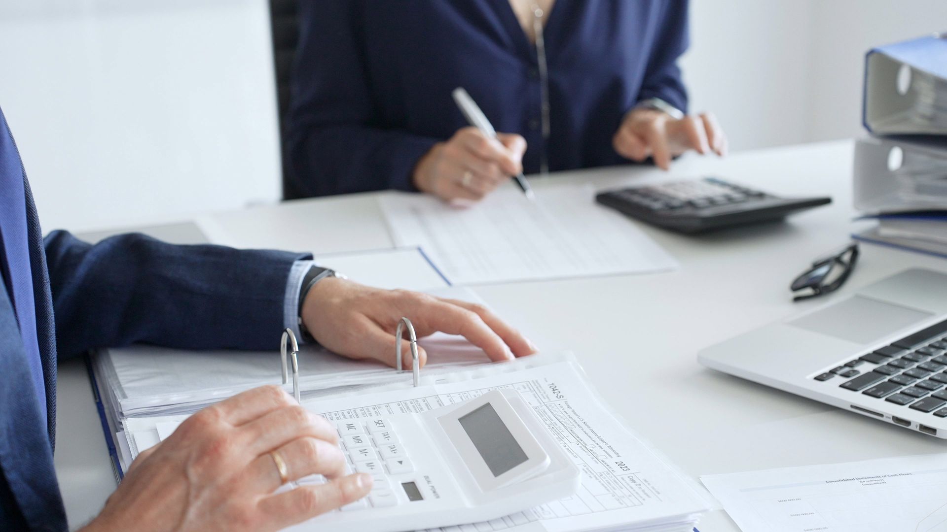 Two people in suits working at a desk with papers, calculators, and a laptop.