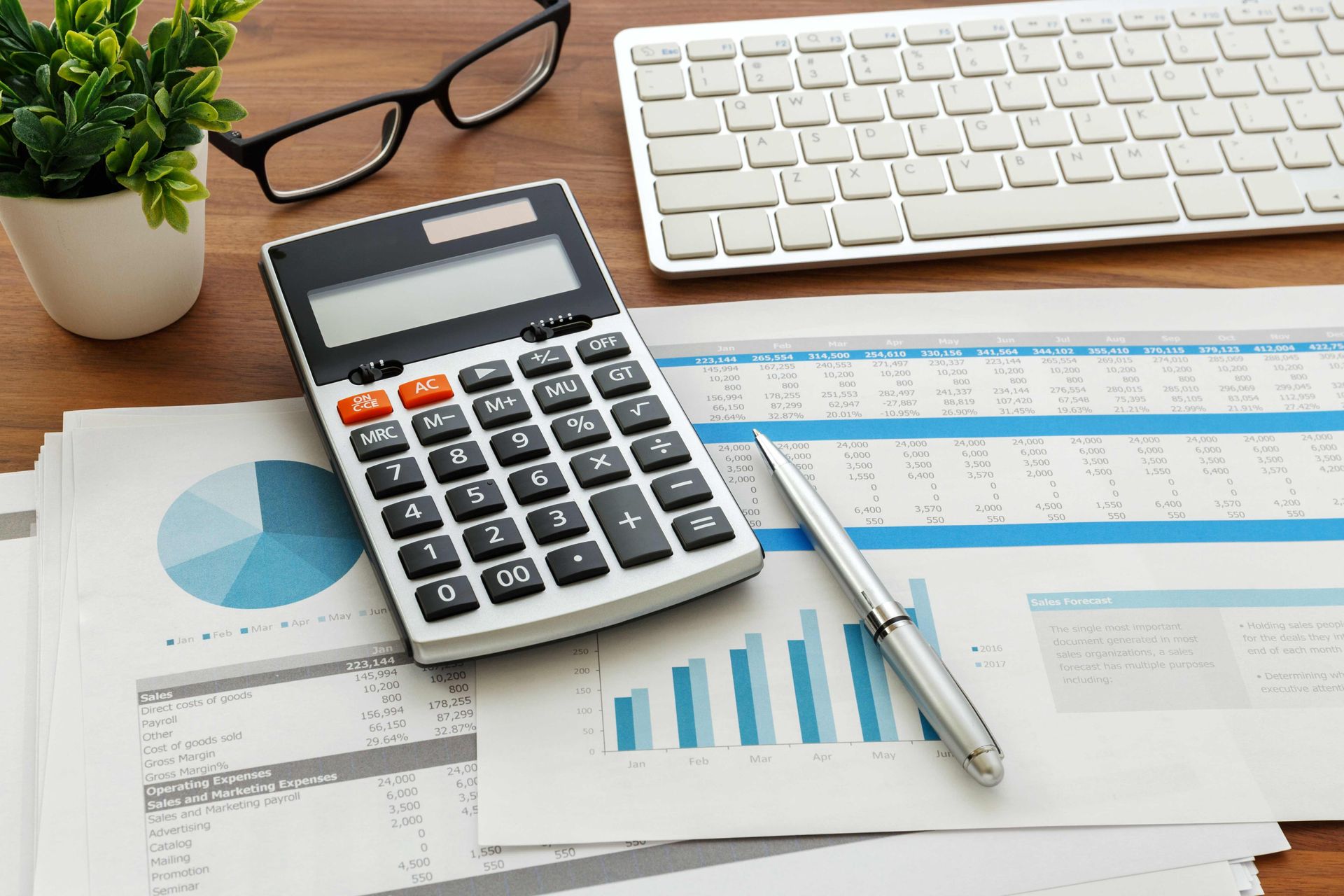 Calculator on a desk with financial charts, keyboard, pen, glasses, and a small plant. Calculator on a desk with financial charts, keyboard, pen, glasses, and a small plant.