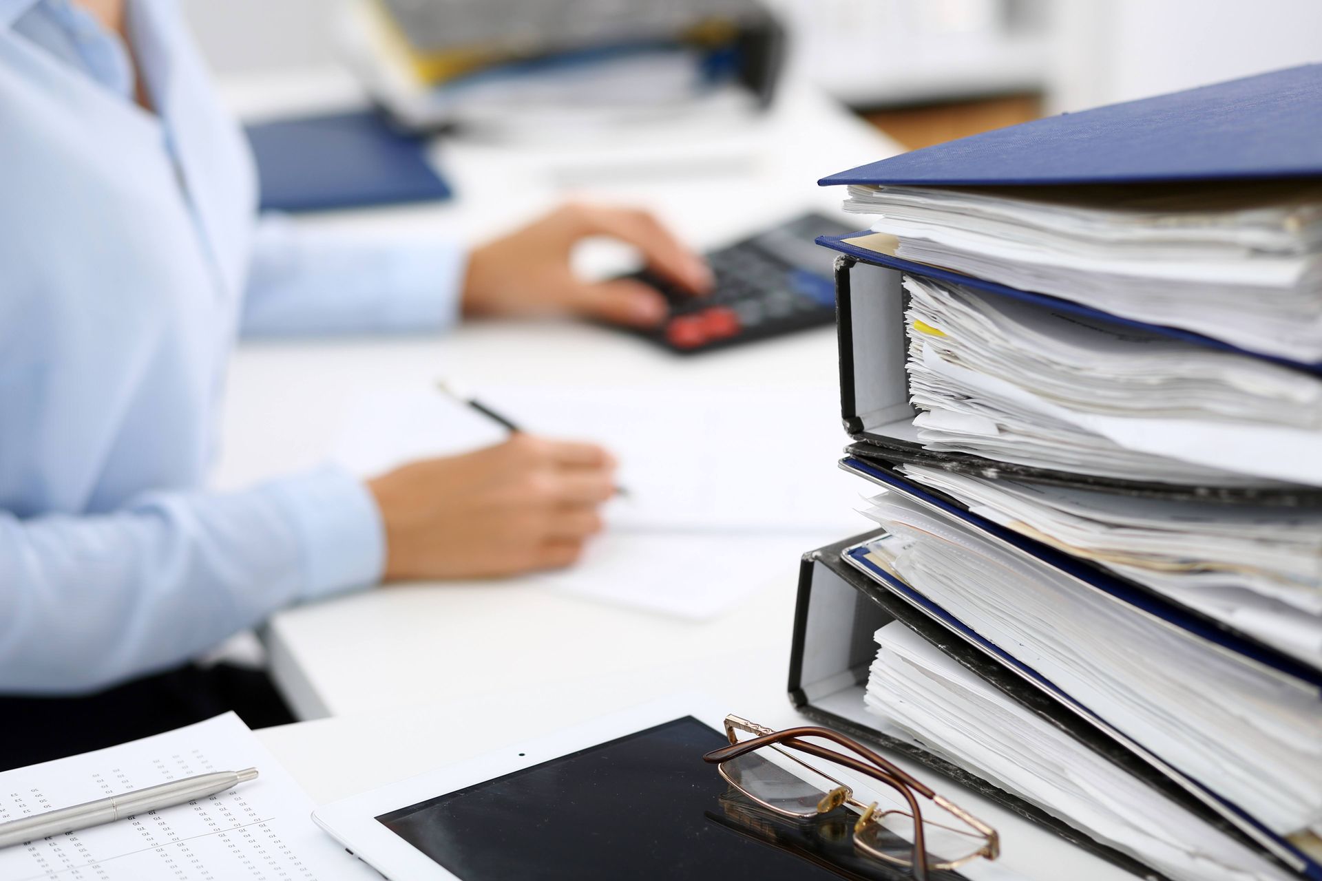Stack of files next to person writing and using a calculator at a desk.