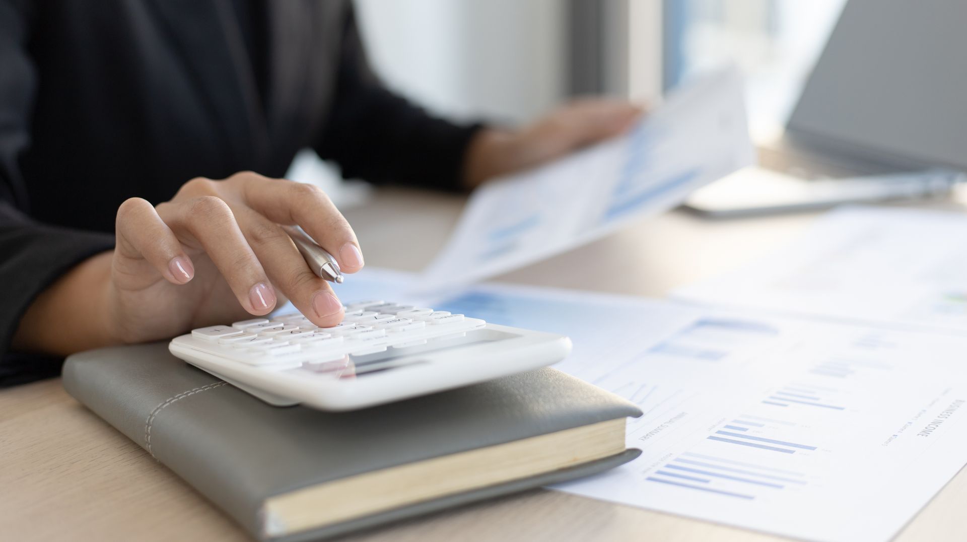 Person's hand using a calculator, resting on a notebook. Papers and a laptop are nearby.