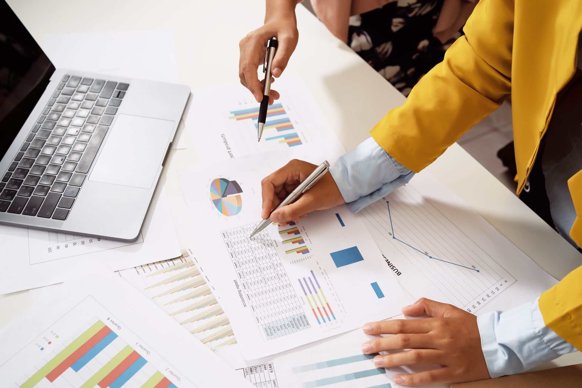 Laptop displays graphs; business analysis documents, pen, and keyboard on a desk.