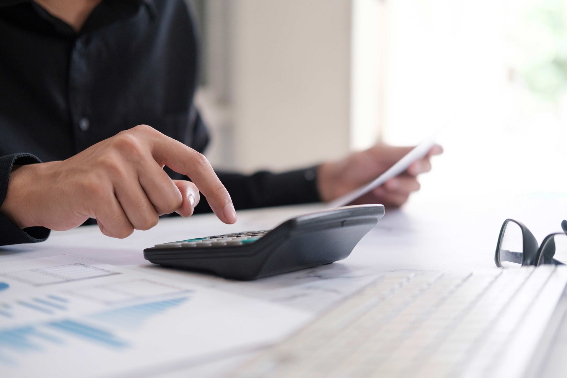 Person using a calculator, holding papers, with glasses on a desk; financial analysis concept.