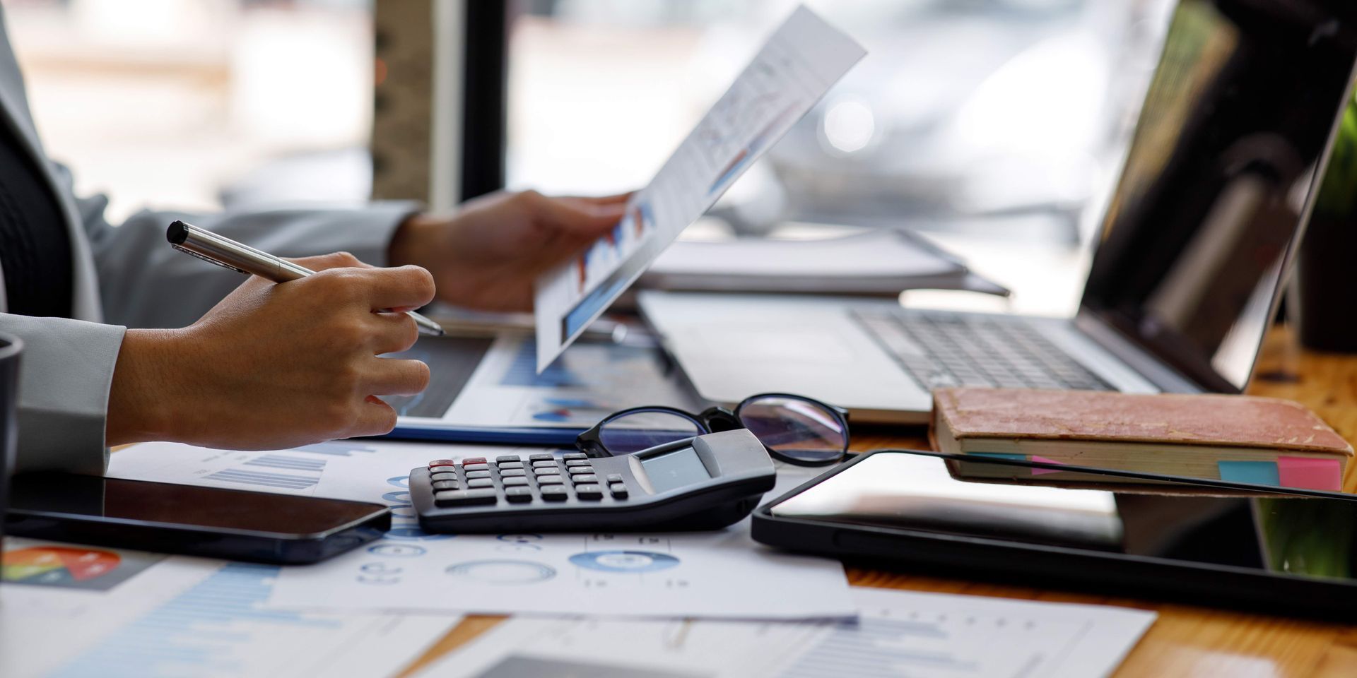 Person reviews documents at a desk, with a laptop, calculator, and smartphone nearby.