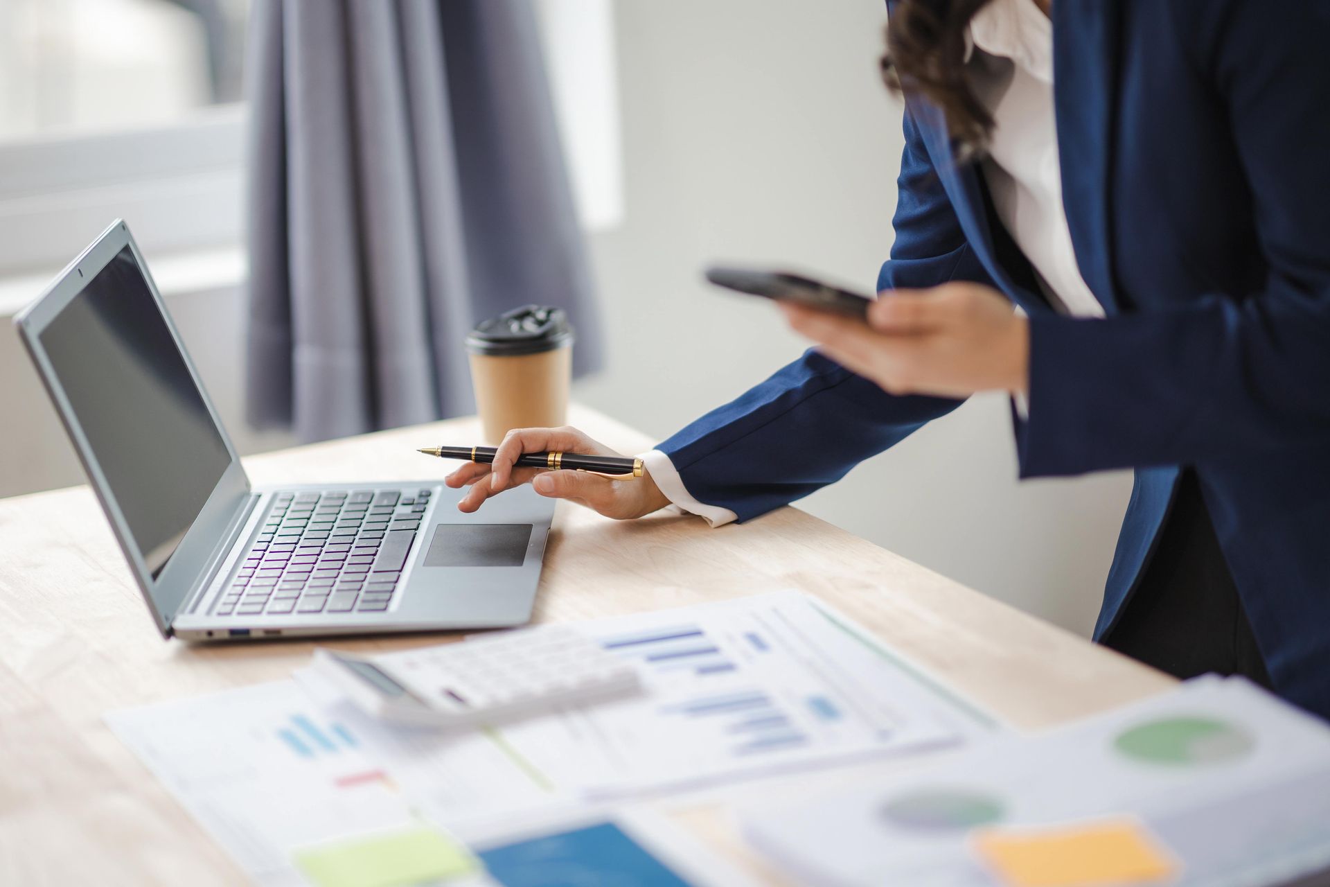 Person in a blue blazer working on a laptop, holding a phone, with financial documents and coffee on a desk.