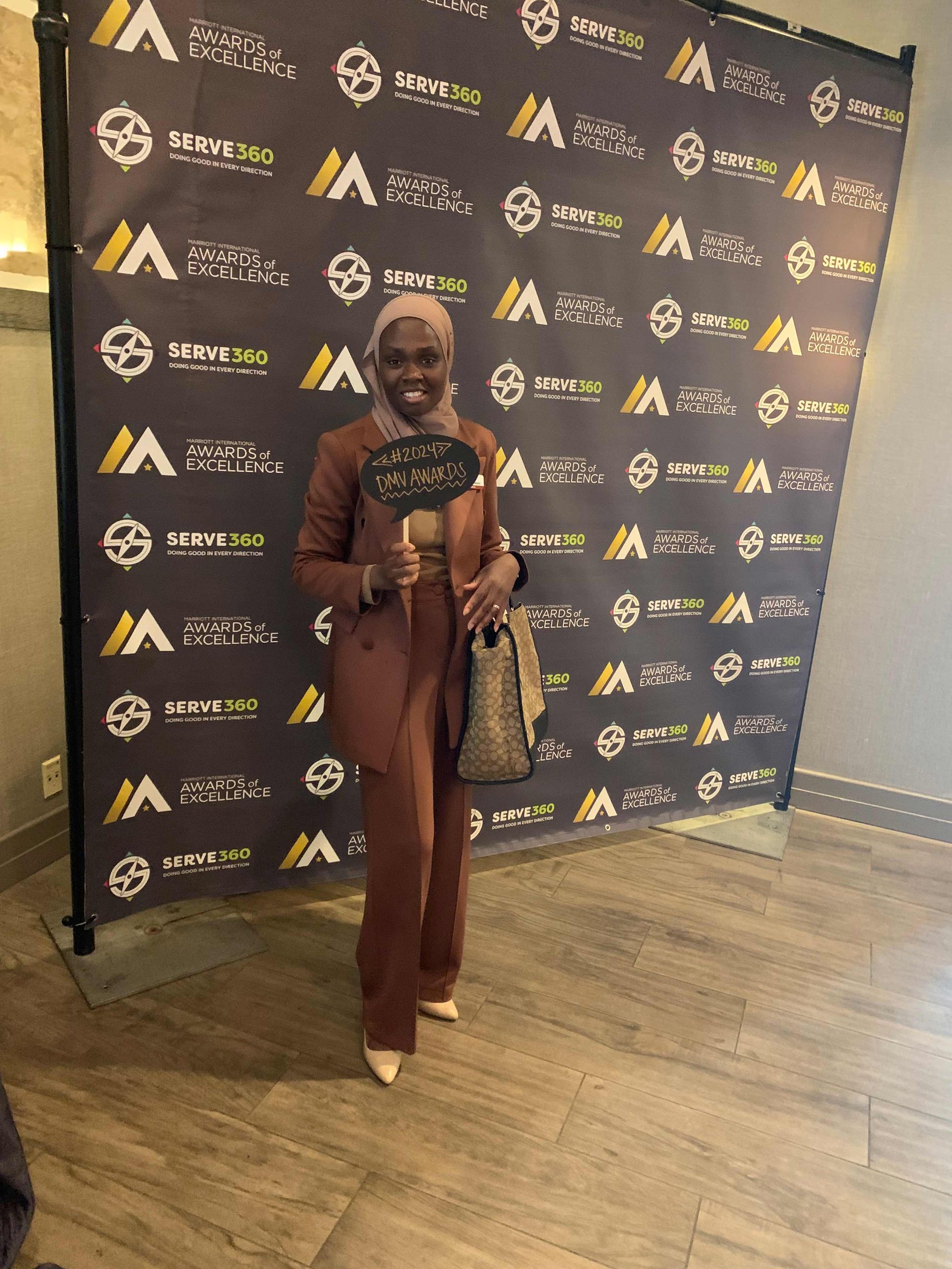 Woman in brown suit poses by a branded backdrop, holding a sign. Woman in brown suit poses by a branded backdrop, holding a sign.