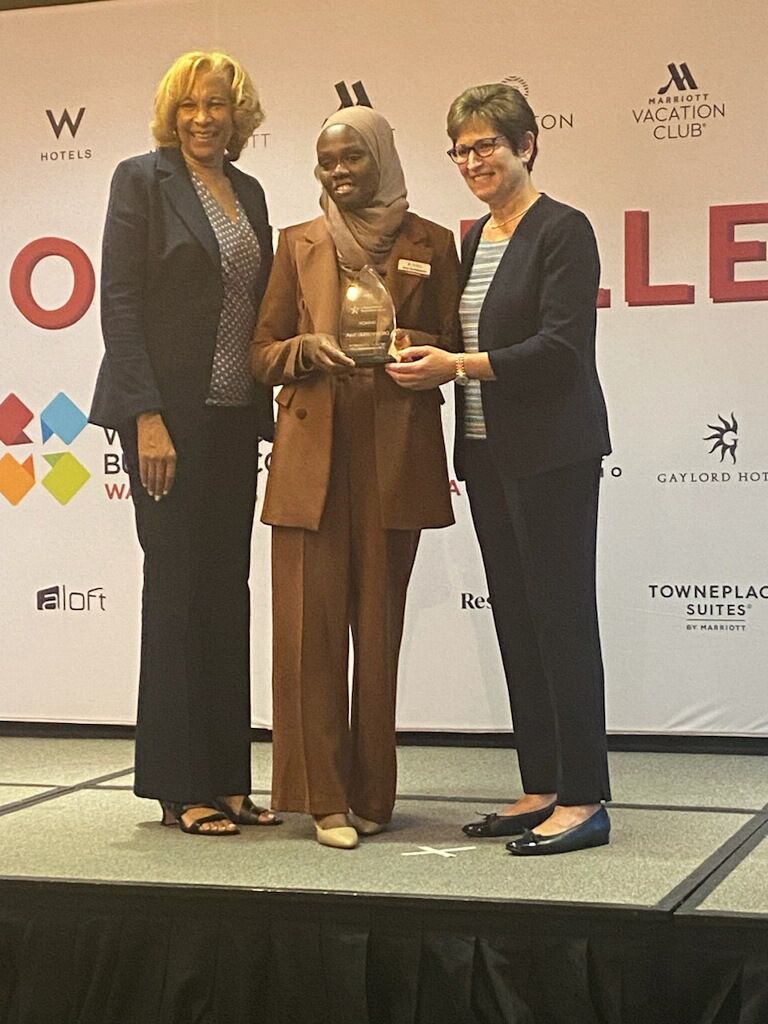 Three people on a stage. One in brown suit holds a trophy, being presented by a person in a black jacket. The woman on the left is in a black pantsuit. Three people on a stage. One in brown suit holds a trophy, being presented by a person in a black jacket. The woman on the left is in a black pantsuit.